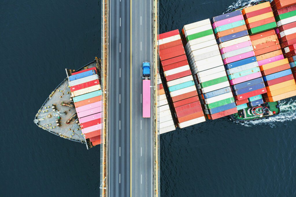 Aerial view of a container ship passing beneath a suspension bridge. Semi truck with pink cargo container crosses above.