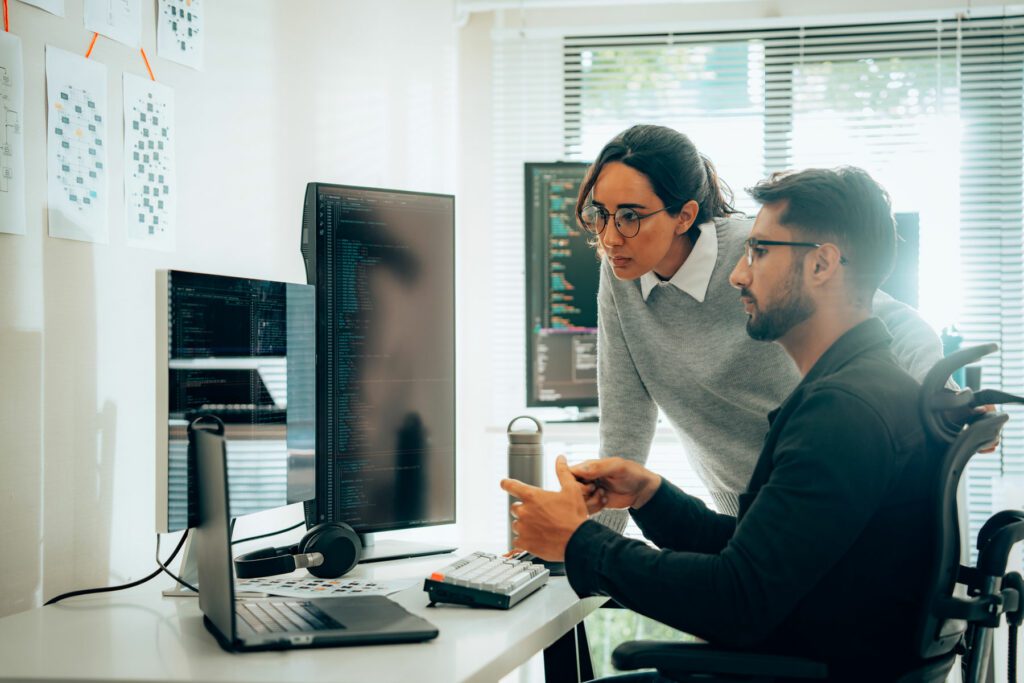 Two technology professionals work diligently in a bright office setting, one focusing on a laptop while the other engages with a large monitor